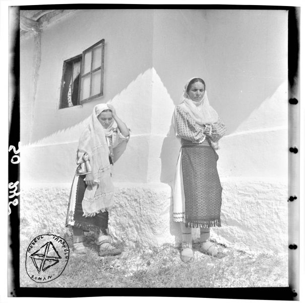 Femei din Oltenia (BA-543) - Aurel Bau(c)h, 01 ian 1938 - 31 dec 1947, Oltenia - În perioada interbelică, în satele olteneşti, coexistau două tipuri de costum tradiţional: intra-, respectiv extracarpatic. Femeia din stânga este îmbrăcată în portul mocanilor, stabiliţi în zonă în urma transhumanţei din părţile Sibiului. Ea poartă cămaşă cu râuri negre, și două şorţuri cu alesătură din fir metalic, peste poale. Femeia din dreapta este îmbrăcată în portul oltenesc, cu o cămaşă cu mâneci cusute „în table”, şi şorţuri ţesute cu acelaşi motiv geometric. Amândouă sunt învelite cu maramă de borangic şi poartă opincile peste ciorapi tricotaţi din lână - foto preluat de pe www.facebook.com/@MuzeulTaranului