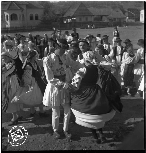 La horă (BA-579) - Aurel Bau(c)h, 10 aug 1945 - 30 noi 1947, Transilvania - Tinerii joacă îmbrăcați în hainele cele bune: femeile poartă cămăși încrețite la gât, cu altiță și râuri pe mâneci, două rânduri de poale, fuste crețe și șorț din pânză industrială și vestă din catifea, iar bărbații poartă cămăși cu poale plisate la cald, pantaloni de lână, chimir de piele, cravată scurtă și pălărie din pâslă groasă. Imagine realizată în cadrul Școlii Sociologice de la București, cu ocazia campaniilor monografice din Valea Gurghiului, desfășurate în anii 1945, respectiv 1947 - foto preluat de pe www.facebook.com/@MuzeulTaranului