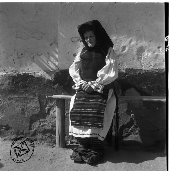 Bătrână în costum de sărbătoare (BA-530) - Aurel Bau(c)h, 01 ian 1938 - 31 dec 1947, Transilvania - Femeia este îmbrăcată în port țărănesc specific zonei Banatului - pieptar înfundat, purtat peste cămașa cu mâneci largi, strânse în manșete cu fodori; poale albe acoperite cu două catrințe din lână (opregul din față este țesut în două ițe, cu decor realizat în năvădeală, iar cel din spate este cu franjuri); și este încălțată în opinci cu gurgui (vârf ridicat), purtate peste obiele colorate din lână fină. Pe cap poartă o legătură neagră, pe suporți - foto preluat de pe www.facebook.com/@MuzeulTaranului