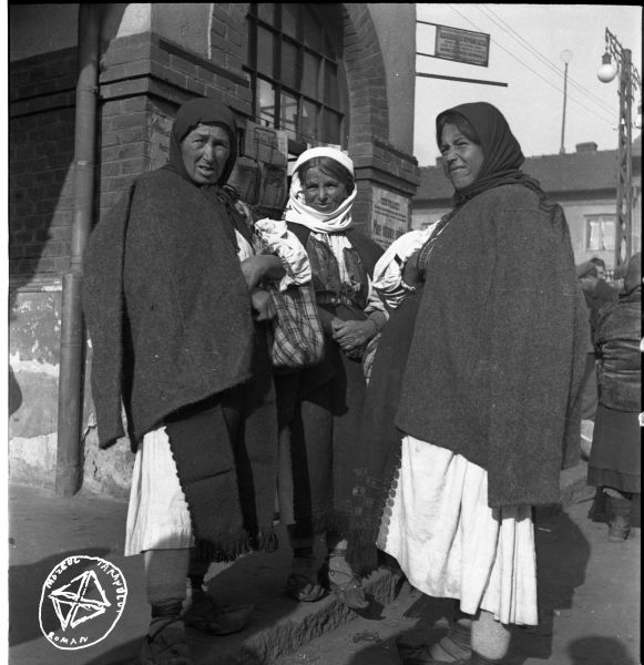 Femei așteptând (BA-573) - Aurel Bau(c)h, 01 ian 1938 - 31 dec 1947, Transilvania Femeile sunt îmbrăcate în portul specific Văii Jiului: cămăși încrețite cu mâneci cu fodori și pieptare despicate în față, brodate pe dinainți, și acoperite de o haină din dimie neornamentată. Au șorțuri negre din lână cu franjuri și dantelă neagră, iar pe cap poartă basmale negre, respectiv velitoare, din ștergar alb legată peste „coarne” - foto preluat de pe www.facebook.com/@MuzeulTaranului